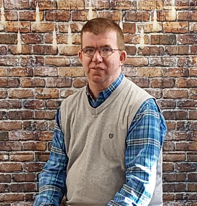 41b178c6-9cb8-ee11-bea1-000d3a54aa76 1 Man in glasses and a gray vest over a blue plaid shirt sitting in front of a brick wall backdrop.