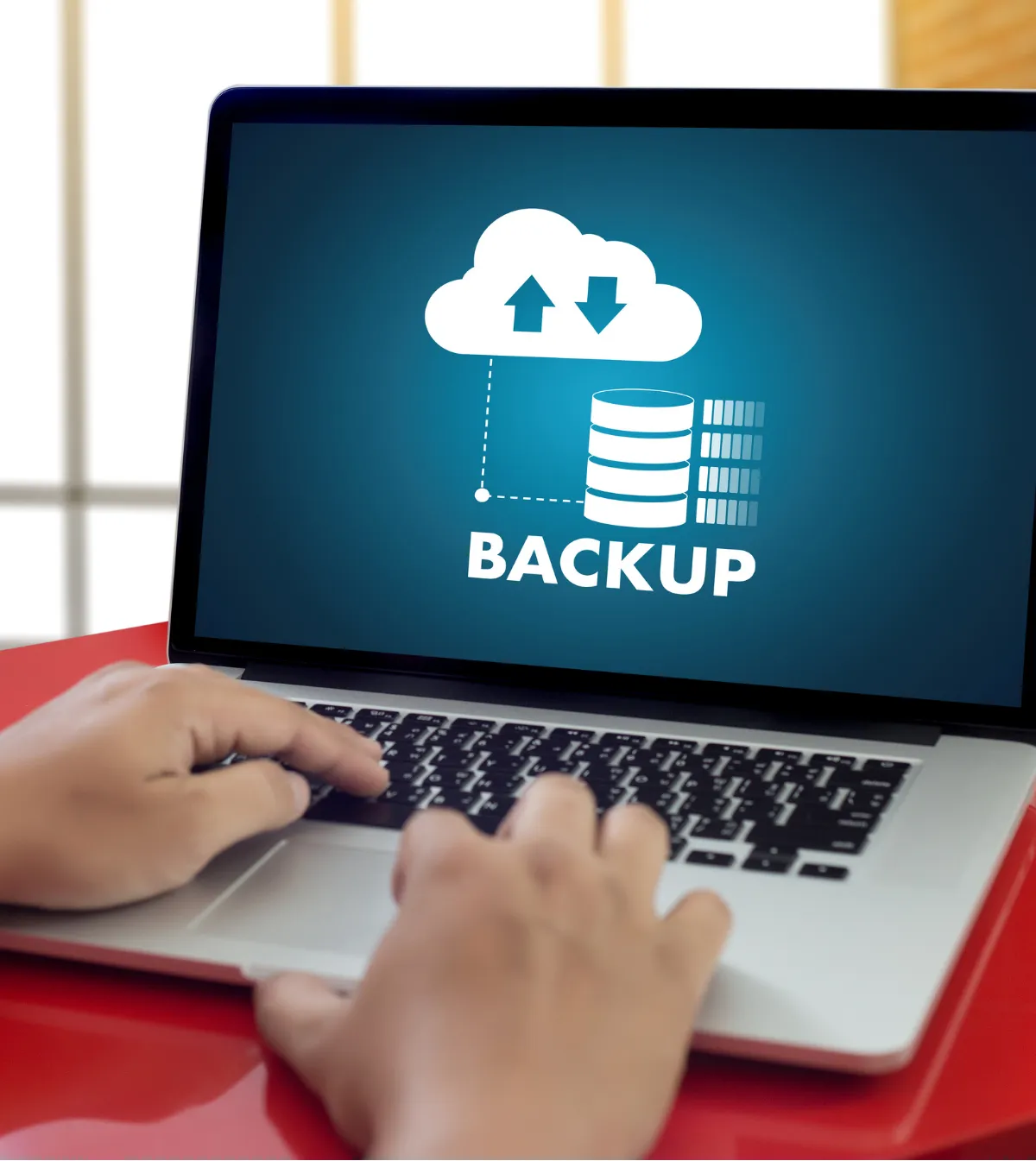 Hands typing on a laptop with a “BACKUP” cloud and database icon displayed on the screen.