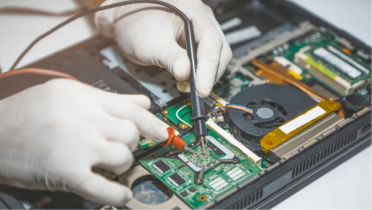 Gloved hands repairing a laptop motherboard with electronic testing tools.