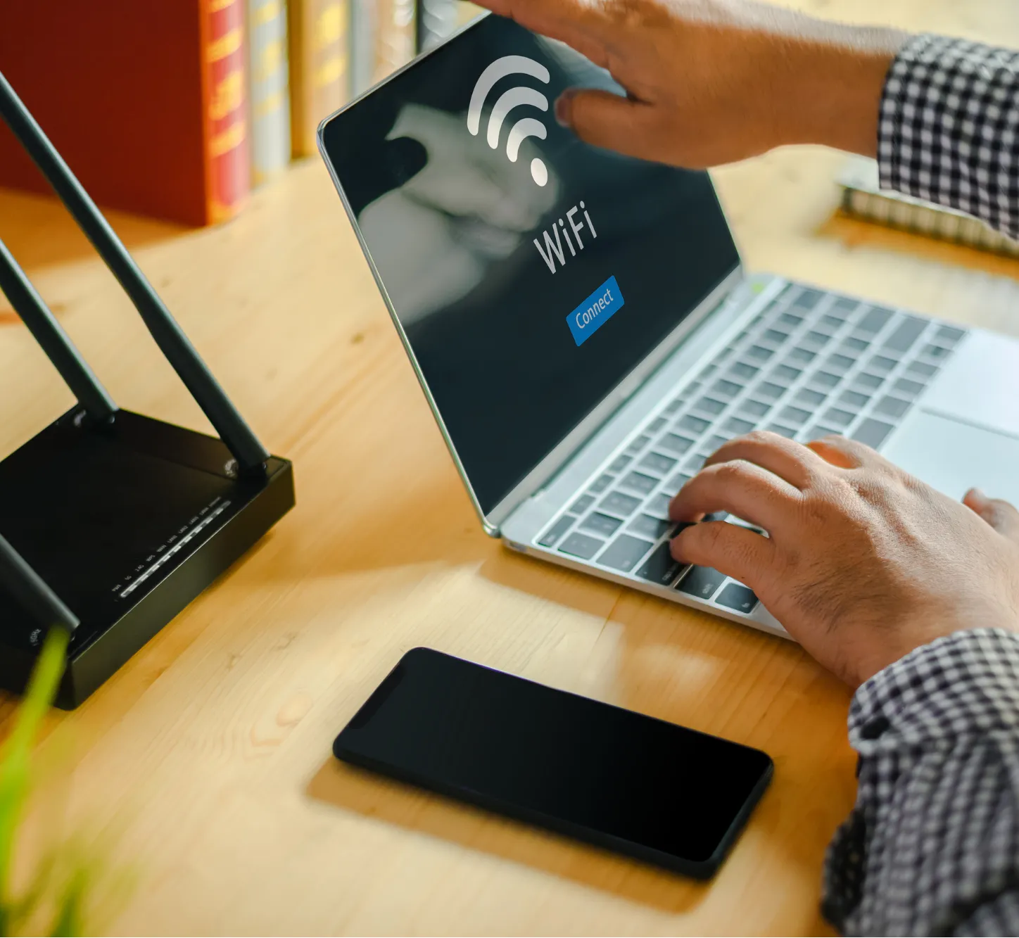 home-office-1 Person connecting a laptop to WiFi, with a phone and a wireless router on a wooden desk.
