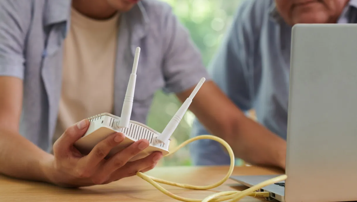 Two people set up a wireless router connected by a yellow cable next to a laptop on a table.