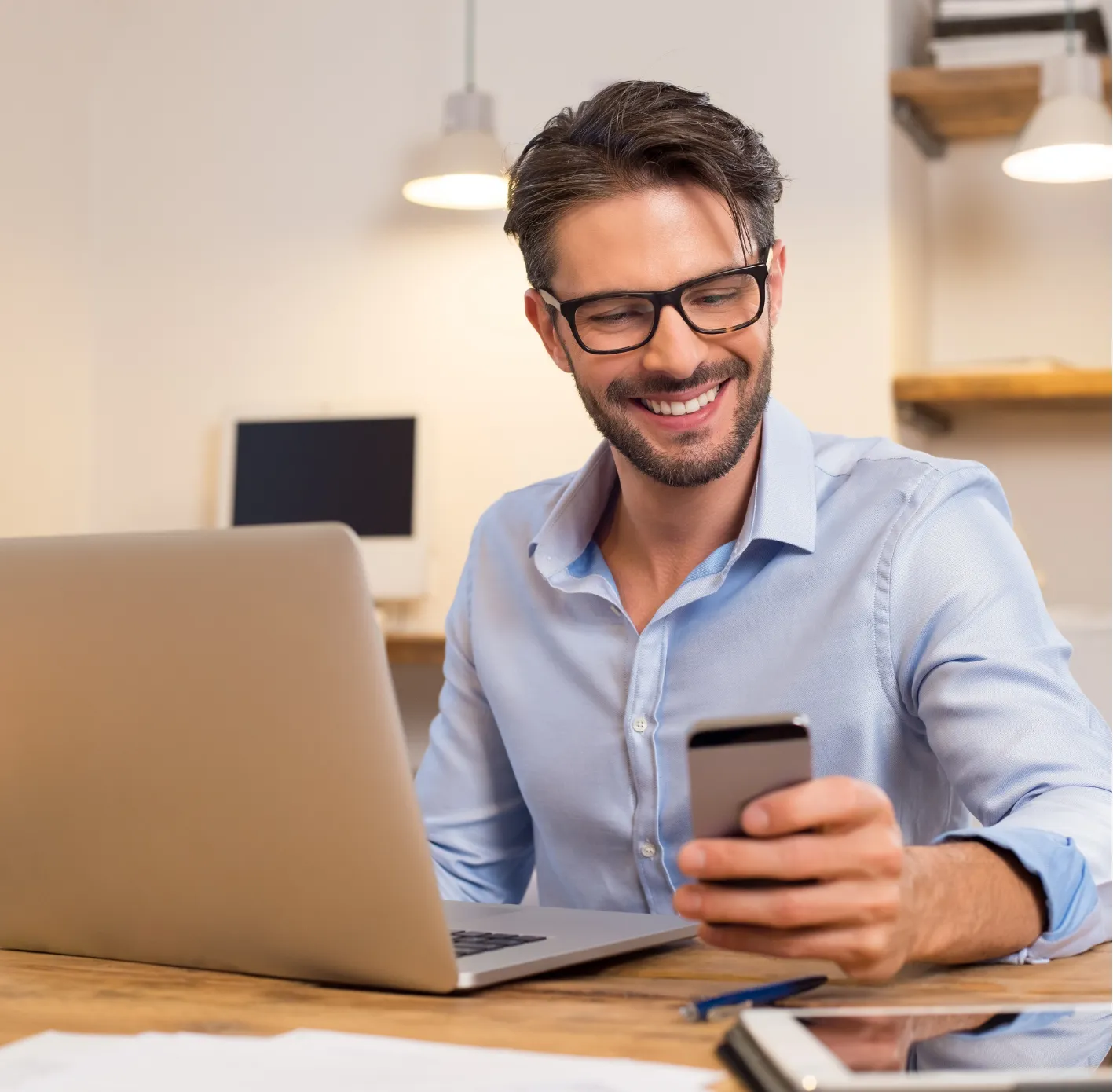 internet-2 Smiling man with glasses looks at his phone while sitting at a desk with a laptop.