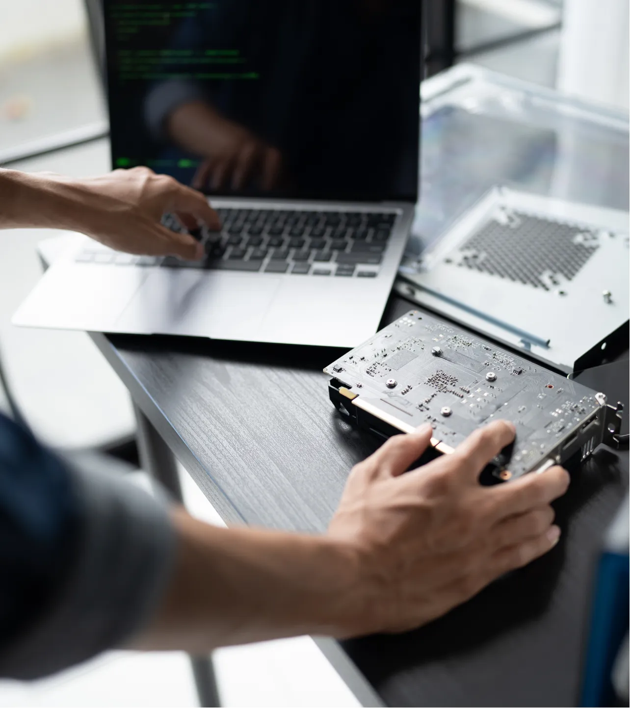 maintenance-2 Person holding a computer part near a laptop with code on the screen, working at a desk.