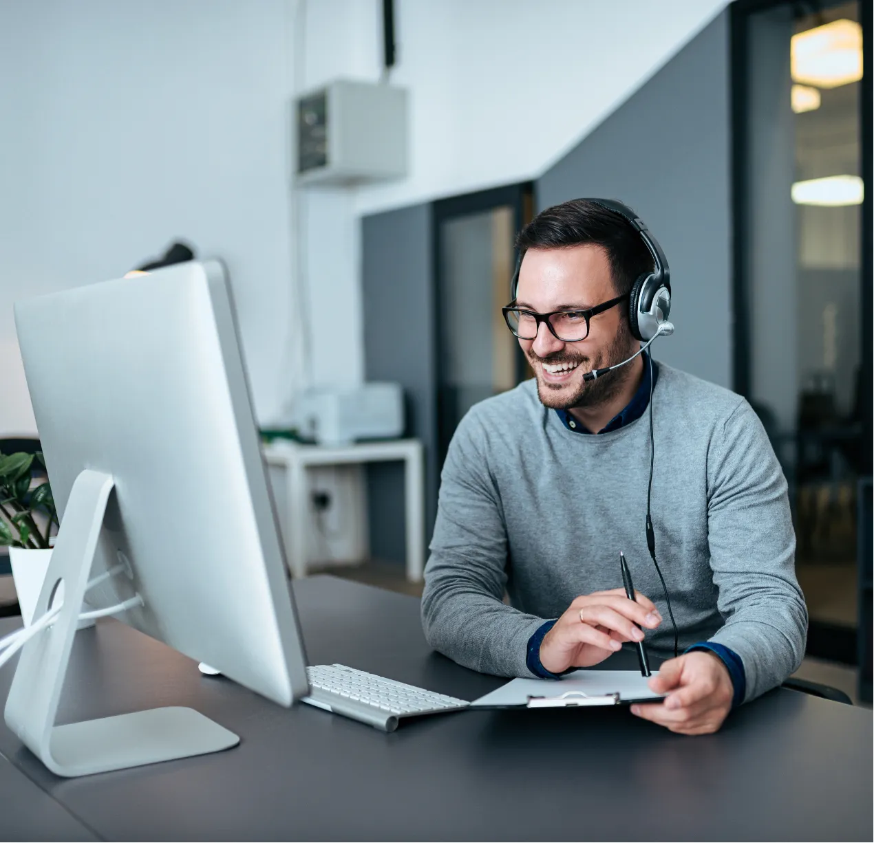 managed-it-1 Man wearing headset, smiling, and taking notes while on a video call at an office desk with a computer.