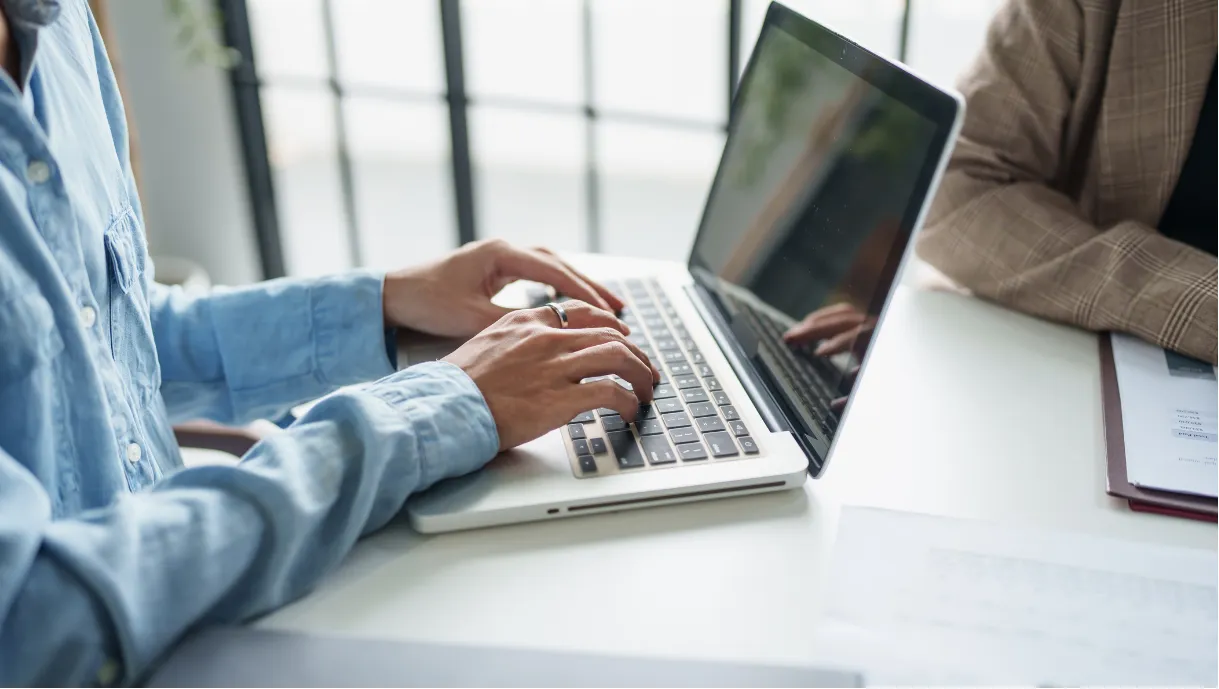 Person typing on a laptop at a desk, with another person sitting nearby, partially visible.