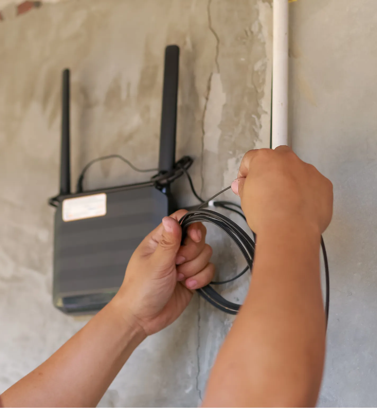 residential-it-2 Hands installing a black internet router with antennas on a wall, adjusting cables next to a white conduit.