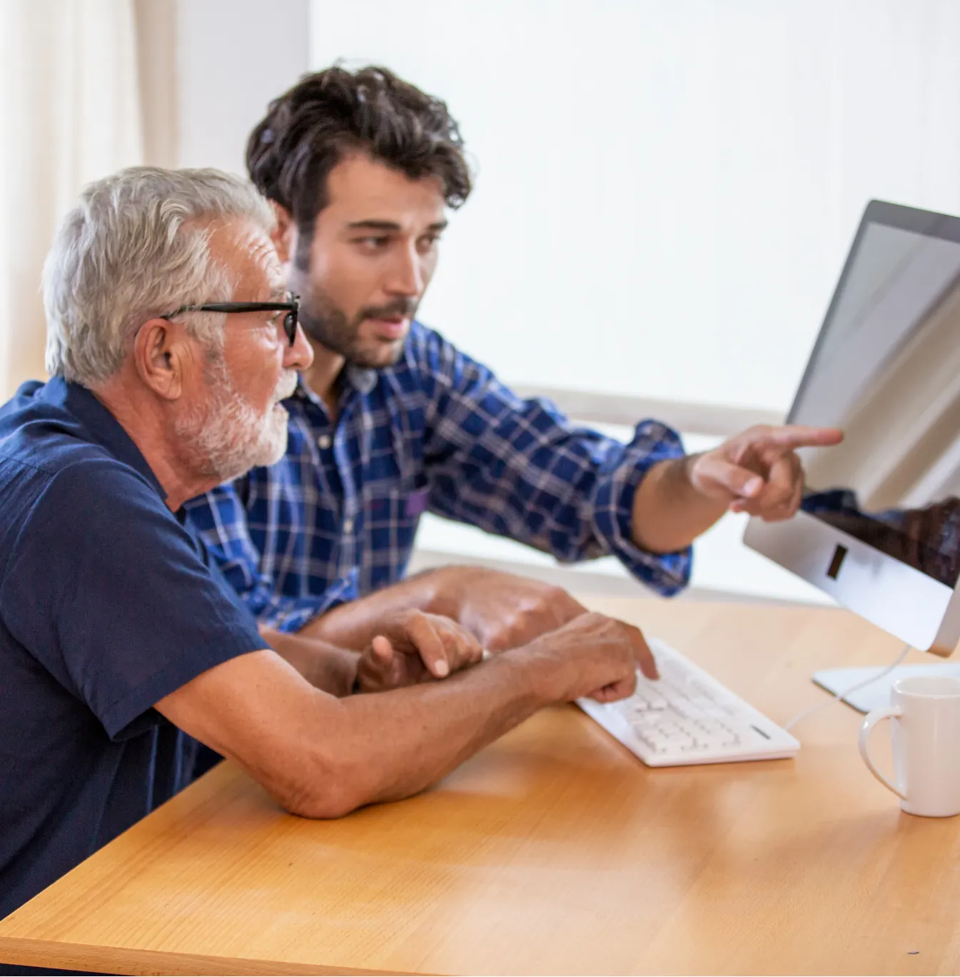 senior-2 Two men sit at a desk, one pointing at a computer screen while the other types on the keyboard.