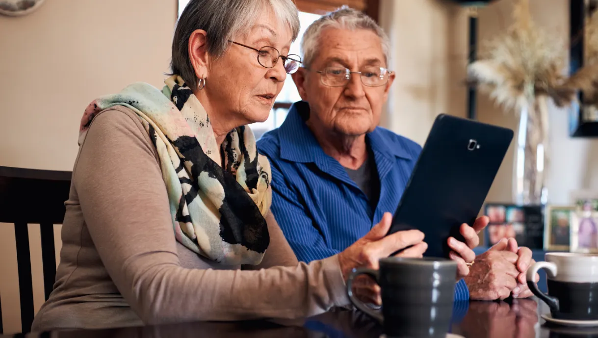 Two older adults sit at a table, looking at a tablet together, with mugs and photos nearby.