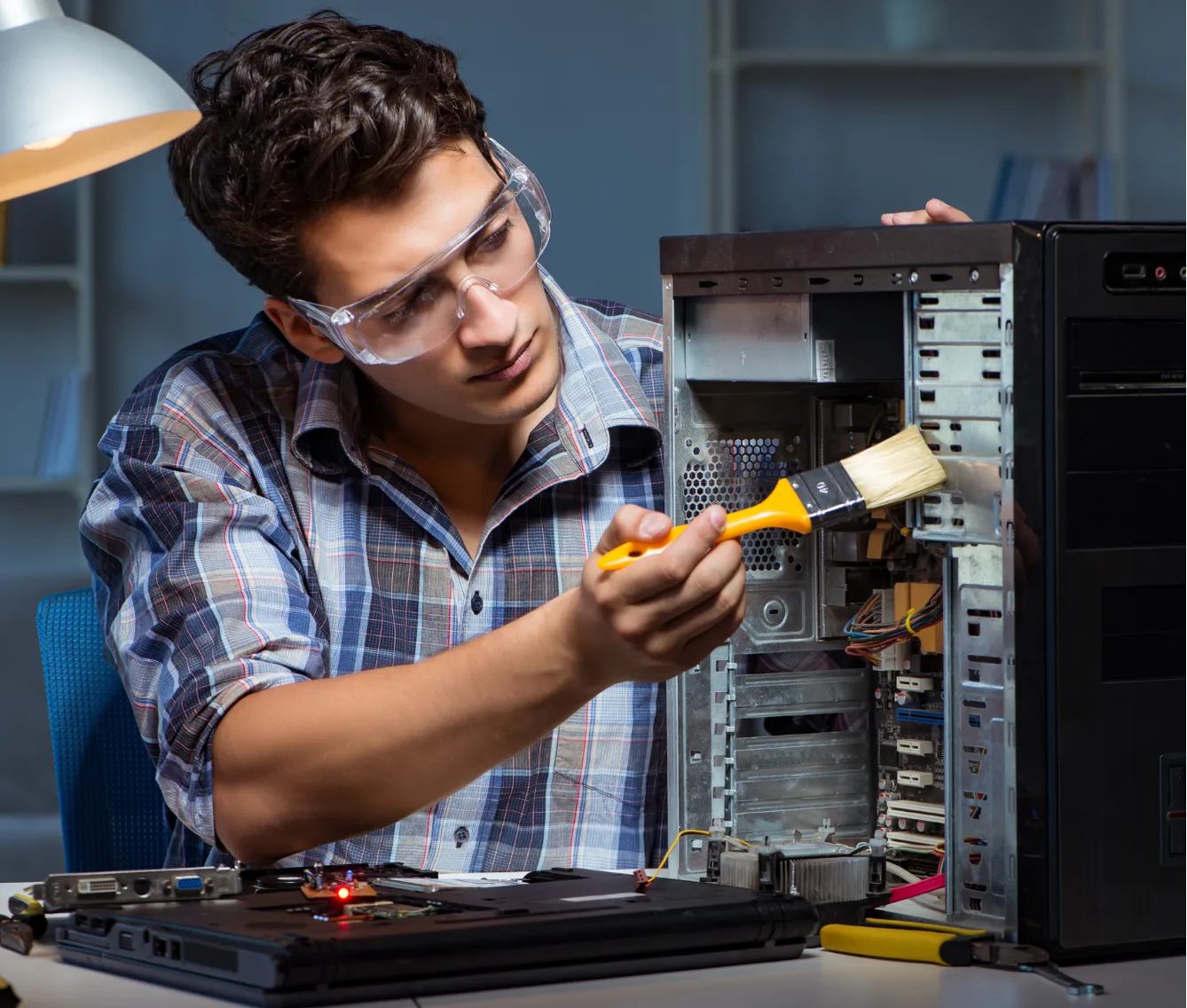 Man wearing safety glasses uses a brush to clean inside a desktop computer tower at a desk.
