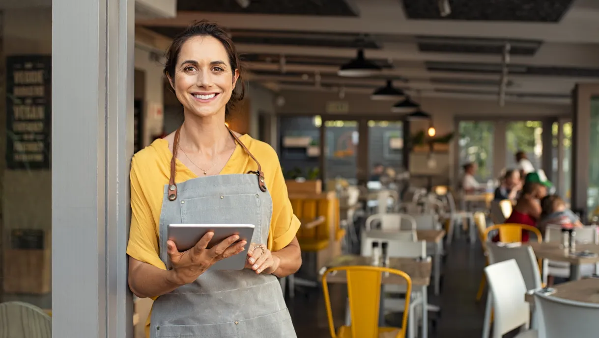 Smiling woman in an apron holding a tablet stands at the entrance of a bright, modern café.