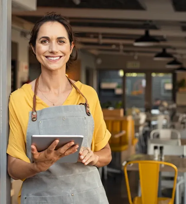 small-business-2 Smiling woman in an apron holds a tablet in a bright, modern café with yellow chairs.