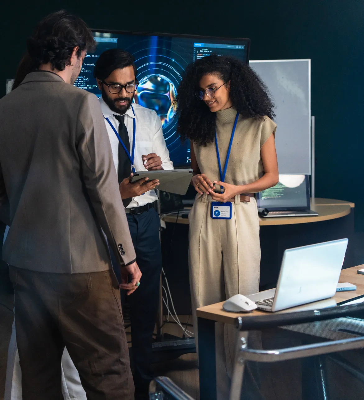 Three people in an office discuss something on a clipboard, with computers and monitors around them.