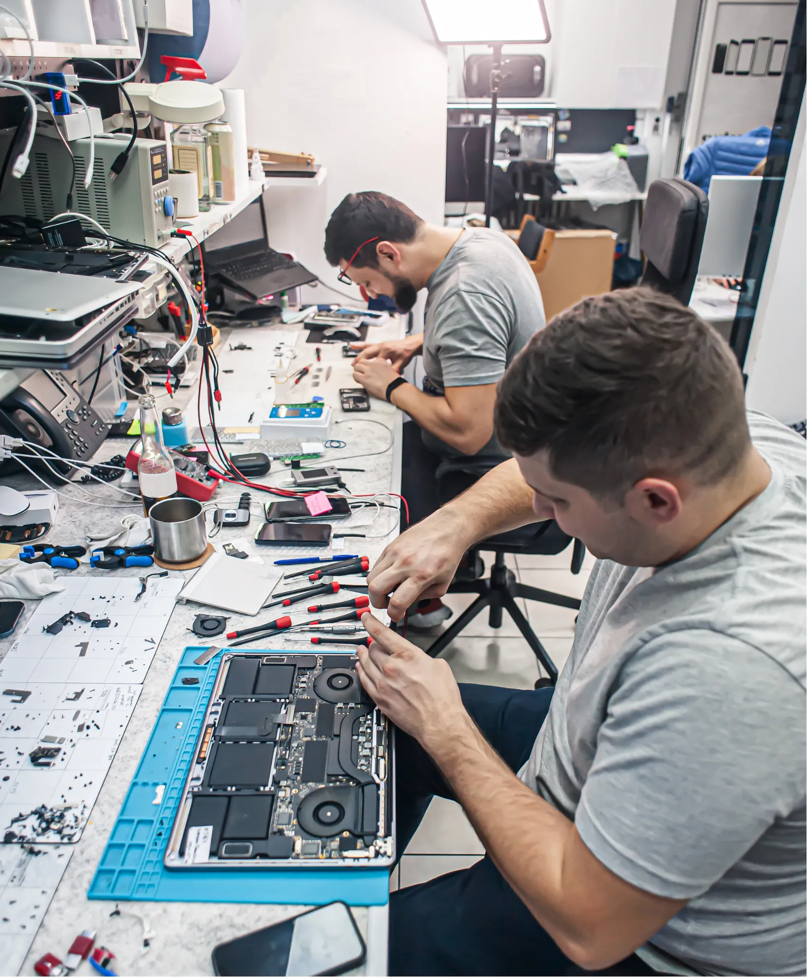 why-s2 Two technicians repair electronics at a cluttered workstation with tools, wires, and open devices.