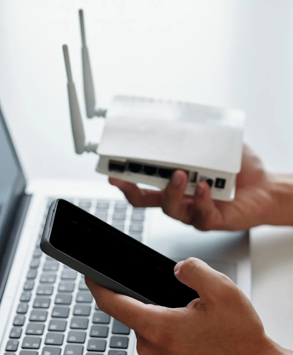 wifi-2 Person holding a Wi-Fi router and a smartphone above a laptop keyboard on a desk.