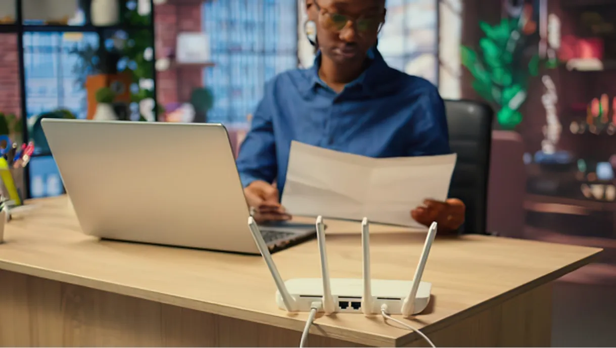 Person at desk with laptop and documents, with a Wi-Fi router in the foreground.