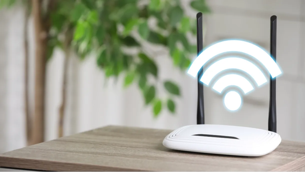A white Wi-Fi router with two antennas sits on a wooden table with a plant in the background.