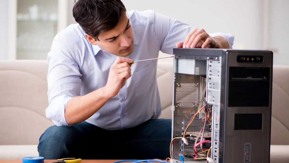 Man fixing a computer tower with a screwdriver, sitting on a couch with tools nearby.